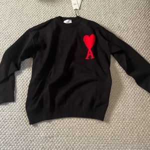 Svart stickad tröja från Ami - Svart stickad tröja från Ami med rund hals och ribbade muddar. På bröstet finns ett stort rött hjärta med bokstaven A, vilket ger en unik och cool detalj. Perfekt för dig som gillar stilrena plagg med en twist.