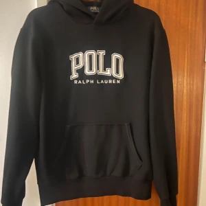 Svart  hoodie från Polo Ralph Lauren - Svart hoodie från Polo Ralph Lauren med stor broderad logga på bröstet och klassisk känguruficka. Tröjan har huva, långa ärmar och liten broderad häst på ärmen. Perfekt för en chill och stilren look.