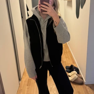 Svart pälsväst med dragkedja - Svart pälsväst med mjuk och fluffig känsla. Västen har en rak passform och stängs med en silvrig dragkedja framtill. Perfekt att slänga över en hoodie för en chill och trendig look.