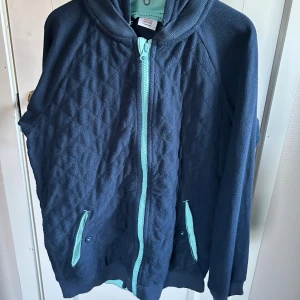 Mörkblå quiltad hoodie med turkosa detaljer - Mysig mörkblå hoodie med quiltad struktur och turkosa detaljer på dragkedja och fickor från PoP. Tröjan har huva, långa ärmar och fickor med knapp framtill. Huvan är löstagbar. Jackan är namnad på insidan (det röda är redigerat på fotot för att maskera namn och nummer. 