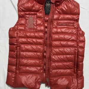 Röd dunväst från Morris, medium - Snygg röd dunväst från Morris i storlek medium. Västen har quiltad design, dragkedja framtill och broderad logga på bröstet. Flera praktiska fickor och en stilren look som passar perfekt till lager-på-lager. Materialet är glansigt och lätt, vilket gör den smidig att bära. Köptes för 300 och är i top skick men lite för kort för mig som är 188cm 