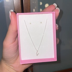 Silverhalsband med V-hänge och stenar - Snyggt halsband i silver med ett V-format hänge dekorerat med små glittrande stenar. Kedjan är tunn och elegant, perfekt för att ge en stilren touch till din outfit. Levereras i en rosa ask.