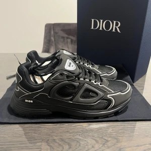 Svarta Dior B30 - Säljer ett par svarta Dior sneakers med vita kontrastdetaljer och chunky sula. Skorna har snörning, meshpaneler och stilrena logotyper på sidan och plösen. Materialmix av skinn och syntet ger en lyxig känsla och modern look. Perfekt för dig som gillar exklusiva streetwear-vibbar.