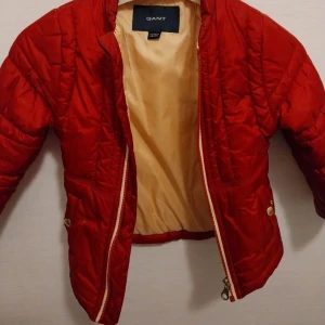 Röd quiltad pufferjacka från GANT - Snygg röd pufferjacka från GANT med quiltad design och guldfärgade knappar på ärmslut och fickor. Jackan har dragkedja framtill och är fodrad i beige. Perfekt för dig som vill ha en cool och varm jacka med detaljer som sticker ut.