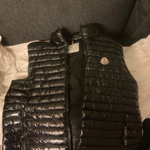 Svart väst från Moncler - .Svart, glansig väst från Moncler, med quiltad design och hög krage. Västen har två sidfickor med dragkedja och Moncler-logotyp på alla dragkedjor. Perfekt för dig som vill ha en elegant och varm look.