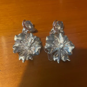Silvriga blommiga örhängen - Stora statement örhängen i silverfärg med blommig design och glittrande sten i mitten. Perfekta för att lyfta din outfit med en cool och unik touch. Blommorna har en snygg, vågig form som ger extra dimension. Orginalpriset låg mellan 200-300kr och eftersom de knappt är använda ligger priset på 100