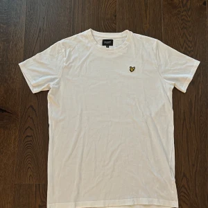 Vit t-shirt från Lyle & Scott Junior - Vit t-shirt från Lyle & Scott Junior med klassisk rund hals och korta ärmar. På bröstet sitter den ikoniska broderade gula örnen. Tillverkad i mjuk bomull för skön känsla och enkel stil. Perfekt för dig som gillar cleana och stilrena plagg.