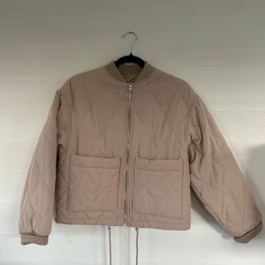 Beige quiltad jacka från Monki - Quiltad beige jacka från Monki i storlek XS. Jackan har stora fickor framtill, dragkedja med rund detalj och ribbade muddar vid hals och ärmslut. Croppad modell med dragsko nertill och snyggt rutmönster över hela jackan.🤩💕