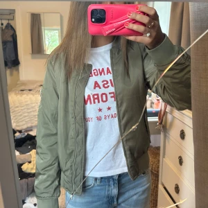 grön bomberjacka  - Snygg olivgrön bomberjacka med klassisk passform och dragkedja framtill. Jackan har ribbade muddar vid ärmslut och midja samt en ärmficka med dragkedja. Perfekt för dig som gillar en stilren och avslappnad look.