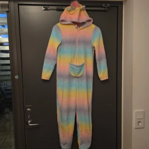 Pastellfärgad fleece hoodie onepiece - Supermysig onepiece i fleece med pastellfärgade ränder i rosa, blått, gult och lila. Har huva med enhörningsdetaljer, dragkedja framtill och en magficka. Perfekt för chilliga dagar hemma.
