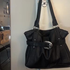 Svart handväska med silverdetalj - Snygg svart handväska med breda axelband och coolt bälte med stor silverfärgad spänne framtill. Perfekt storlek för att få plats med allt du behöver, kommer tyvärr inte till användning💕💕