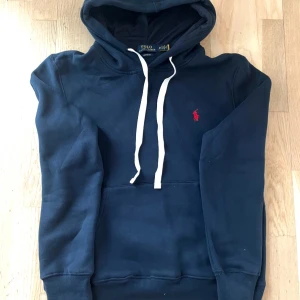 Marinblå hoodie från Polo Ralph Lauren - Säljer en marinblå hoodie från Polo Ralph Lauren med vit snörning och klassisk röd logga på bröstet. Tröjan har huva, känguruficka och är tillverkad i mjuk bomull. Perfekt för dig som gillar stilren och bekväm streetwear.