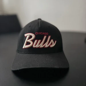 Chicago Bulls keps från Mitchell & Ness - Svart keps med böjd skärm och broderad Chicago Bulls-logga i vitt och rött framtill. Sidan har Bulls-symbolen i rött och vitt, och baktill står Mitchell & Ness broderat. Justerbar snapback. Perfekt för dig som gillar basket och streetwear.