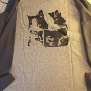 Grå långärmad tröja med kattmotiv (helt nya) - Grå långärmad tröja med svart tryck av fyra kattansikten på bröstet. Materialet är mjukt och har en diskret struktur. Perfekt för dig som gillar djur och vill ha en chill och unik stil. Ärmarna är i mörkare grå ton och tröjan har en relaxed passform.