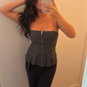 Mörkgrå korsett-topp med dragkedja - Säljer en mörkgrå korsett-topp med axelbandslös design och markerad midja. Toppen har en synlig dragkedja framtill och två stora fickdetaljer nedtill. Lägger sig perfekt runt kroppen så man får en snygg figur. Varsamt använd. Otrolig snygg men behöver nytt hem! Jag är vanligtvis S så den är liten i storleken 