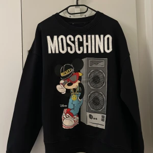 Svart Moschino x Disney sweatshirt H&M  - Svart sweatshirt från H&M i samarbete med Disney och Moschino. Stor broderad MOSCHINO-text och coolt tryck med Mickey Mouse i streetstyle framför en högtalare. Långärmad, rund hals och ribbade muddar. Perfekt statement-plagg för dig som gillar unika prints. Storlek L, Kragtaggen har klippts av