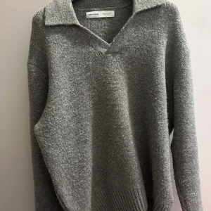 Grå stickad tröja från Bershka - Mysig grå stickad tröja från Bershka med v-ringning och krage. Tröjan har lång ärm och ribbade muddar vid ärmslut och nederkant. Perfekt för lager-på-lager och chill höst- eller vinterstil.