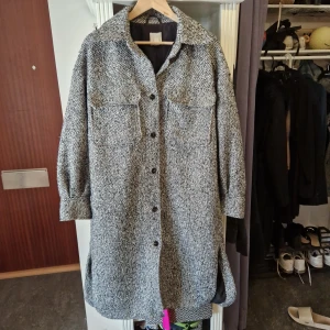 Oversize höstkappa från H&M, gråsvart strl M - Stilren oversize kappa från H&M i gråsvart melerad . Jackan har stora bröstfickor, knappar framtill och klassisk krage. Perfekt för lager-på-lager och har en loose passform med långa ärmar och slitsar i sidorna.