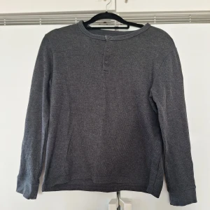 Mörkgrå långärmad tröja med knappar M - En enkel och stilren mörkgrå långärmad tröja med rund halsringning och tre knappar framtill. Tröjan är tillverkad i ett mjukt bomullsmaterial och passar perfekt för lager-på-lager under kyligare dagar.
