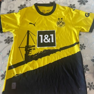 Borussia Dortmund fotbollströja Puma - Gul och svart Borussia Dortmund fotbollströja från Puma med klubbmärke, sponsorlogga och trycket DORTMUND på ryggen. Tröjan har korta ärmar, svart krage och detaljer, samt grafiskt tryck framtill. 