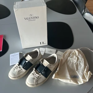 Valentino Garavani Open sneakers vit/svart - Snygga Valentino Garavani Open sneakers i vitt skinn med bred svart läderdetalj över mitten. Klassisk rund tå, platt sula och snörning. Kommer med originalkartong, dustbag och äkthetskort. Perfekta för dig som gillar lyxiga och stilrena sneakers. Kan tyvärr inte garantera äkta men köpta på plick för 1799