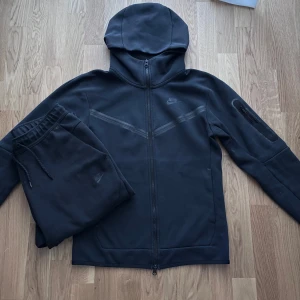 Svart Nike Tech set XS/S - Svart Nike Tech set. Tröjan är storlek XS med hel dragkedja och huva. Tröjan har långa ärmar, diskret Nike-logga på bröstet och en praktisk ficka med dragkedja på ena ärmen. Matchande byxor i storlek S medföljer, båda i mjuk bomullsblandning.