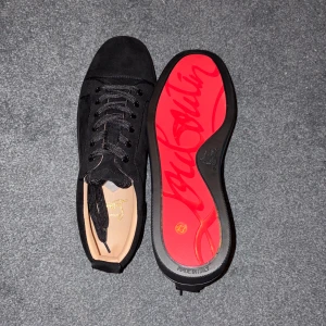 Svarta sneakers från Christian Louboutin - Snygga svarta sneakers från Christian Louboutin i mocka med klassisk röd sula och svarta skosnören. Skorna har rund tå och platt sula, samt diskreta metalldetaljer på sidan. Perfekta för dig som vill sticka ut med exklusiv stil.