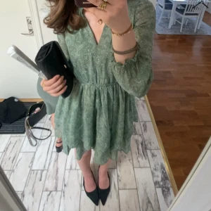 Grön kortklänning med volang - Superfin grön kortklänning med diskret blommigt mönster och volangkant. Klänningen har v-ringning, långa transparenta ärmar och resår i midjan för en snygg passform. Perfekt för dig som gillar en luftig och söt stil.