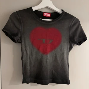 Svart croppad topp med rött hjärta Diesel - Cool svart croppad topp från Diesel med ett stort rött hjärta och diskret logga på bröstet. T-shirten har rund halsringning och korta ärmar. 