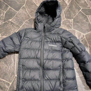 Svart dunjacka Peak Performance stl 160 - Säljer en svart pufferjacka från Peak Performance med huva och dragkedja framtill. Jackan är quiltad och har en klassisk, sportig look. Perfekt för kalla dagar och enkel att matcha med olika outfits.är några små ljusa prickar på en axel annars väldigt bra skick