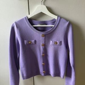 Lila kofta från ELSA.M med guldknappar - Superfin lila kofta från ELSA.M med snygga guldknappar framtill och på axlarna. Koftan har rund halsringning, långa ärmar och två dekorativa fickor på bröstet. Perfekt att slänga över en topp för en trendig look.