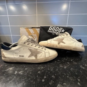 Golden Goose vita sneakers med stjärna - Golden Goose sneakers i vitt skinn med grå mockadetaljer och ikonisk stjärna på sidan. Grå snörning, svart hälparti och platt sula. Klassisk low-top modell med GGDB-logga på sidan. Snyggt slitna detaljer för en cool look. Bedöm skicket själv så inget fel kommer upp eller händer och vid frågor svarar jag snabbt