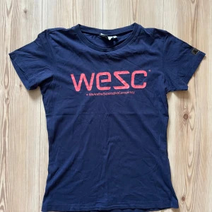 Mörkblå t-shirt från WESC i bomull - M - Snygg mörkblå t-shirt från WESC med röd logga framtill. Klassisk rund halsringning och korta ärmar. Tillverkad i mjuk bomull, perfekt för en avslappnad stil. Passformen är normal och materialet känns skönt mot huden.