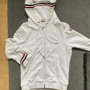 Moncler tröja med dragkedja - Snygg vit hoodie från Moncler med dragkedja, huva och snörning. Klassiska Moncler-ränder i rött, blått samt logga på armen går att scanna! Skriv för mer information👍🏼