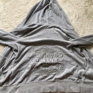 Grå Juicy Couture hoodie med dragkedja Storlek M - Säljer en grå hoodie från Juicy Couture med broderad logga och krona på ryggen. Tröjan har huva, dragkedja framtill och två fickor. Mjuk och skön. Klassisk streetstil med snygga detaljer.
