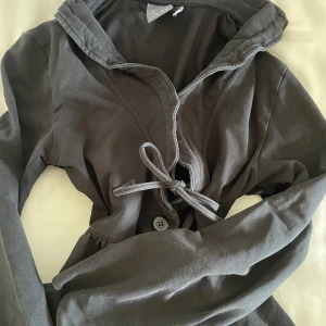 Svart tröja med knyt från House of Lola - Svart tröja från House of Lola i mjuk bomull. Tröjan har långa ärmar och knytband framtill samt knappdetalj. 