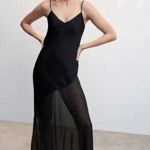 Svart långklänning med skirt material nedtill - Svart långklänning med tunna axelband och v-ringning. Klänningen har en asymmetrisk design med transparent nederdel i lätt, flowy material. Passar perfekt till black tie, sittning och bal! Helt oanvänd med prislapp kvar.