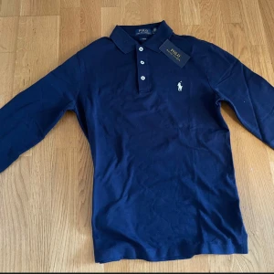 Marinblå pikétröja från Ralph Lauren - Snygg marinblå långärmad pikétröja från Ralph Lauren i slim fit. Klassisk krage, tre vita knappar och broderad logga på bröstet. Tillverkad i mjuk bomull som känns skön mot huden. Pris kan diskuteras. Bara fråga om du undrar något!
