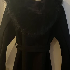 Svart kappa med äkta päls  och bälte - Snygg svart kappa med bred krage i äktapäls  och knytbälte i midjan. Jackan har lång ärm och en stilren, rak siluett som ger en cool vibe. Perfekt för dig som vill ha en elegant look med extra edge under kyliga dagar. Den är lite nopprig men går lätt att få bort säljer den pågrund av att den är förliten kommer ej ihåg vart jag köpt den pris går att diskutera.