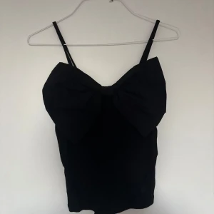 Svart linnetopp med stor rosett - Säljer en svart linnetopp med smala axelband och en stor, iögonfallande rosett framtill. Toppen har en tight passform och är perfekt för dig som vill sticka ut med en cool detalj. Enkel men ändå superunik design.