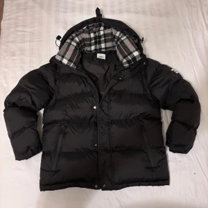 Burberry jacka - Svart pufferjacka från Burberry med avtagbara ärmar, så den kan bäras som väst. Jackan har huva med svartvitt rutigt foder och dragkedja framtill. Tydlig Burberry London England-logga på ärmen och två dragkedjefickor. Perfekt för kalla dagar.