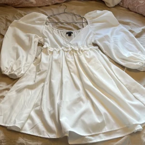Vit omlottklänning från Boohoo - Supersöt vit kortklänning från Boohoo med puffiga långärmar, omlott-skärning och volangdetalj i midjan, oanvänd pga köpte i fel storlek. Perfekt till student, skolavslutning eller bara till vardags. Klänningen har en flowy kjol och elastiska ärmslut för en extra trendig look. Perfekt för dig som gillar romantisk och stilren vibe 🤍