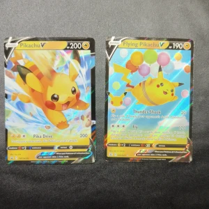 Pikachu V & Flying Pikachu V (Pokémonkort) - Två coola och glittriga Pokémonkort: Pikachu V och Flying Pikachu V! Perfekta för samlare eller dig som vill boosta din lek med ikoniska Pikachu. Korten är i fint skick och har snygg holografisk design som verkligen sticker ut.