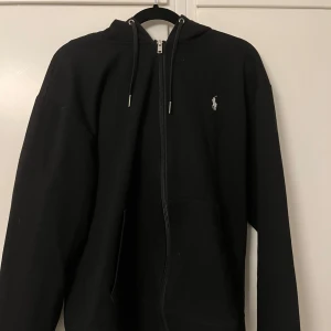 Svart hoodie från Polo Ralph Lauren - Svart hoodie från Polo Ralph Lauren med dragkedja framtill och klassisk vit broderad logga på bröstet. Tröjan har huva med snörning och känguruficka. Perfekt för chill dagar och enkel att matcha med allt. Litet hål i fickan men inget man märker 