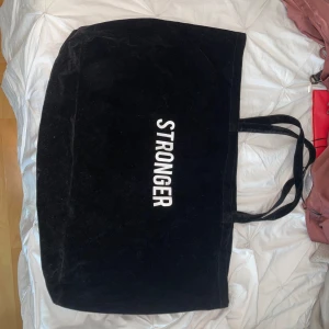 Stor svart träningsväska från Stronger - Säljer en stor svart träningsväska från Stronger med vita bokstäver på framsidan. Väskan har två långa handtag och är tillverkad i mjuk bomull. Perfekt för att bära med sig allt man behöver till skolan, gymmet eller stan.