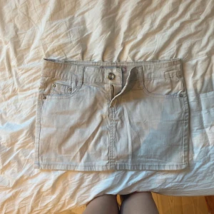 Beige jeanskjol från Miss Azurra - Snygg beige jeanskjol från Miss Azurra med klassisk femficksdesign, bälteshällor och dragkedja framtill. Bakfickorna har broderade detaljer som ger kjolen en extra touch. Perfekt för dig som gillar en enkel men trendig look.