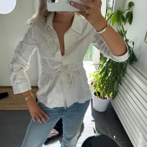 Vit skjorta med knyt och knappar - Snygg vit skjorta i bomull med markerad midja och knytband. Skjortan har klassisk krage, knappar framtill och långa ärmar. Perfekt för dig som gillar en feminin och stilren look.