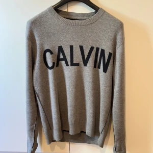 Grå stickad tröja Calvin Klein Jeans - Snygg grå stickad tröja från Calvin Klein Jeans med stor svart CALVIN-text framtill. Rund hals och långa ärmar, perfekt för dig som gillar stilrena och trendiga plagg med tydlig logga. Skön och mjuk kvalitet som passar till många outfits.