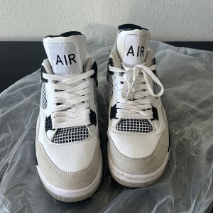 Nike Air Jordan 4 vit/svart/grå - Säljer ett par Nike Air Jordan 4 sneakers i vitt, svart och grått. Skorna har klassisk meshpanel, detaljer i mocka och skinn samt tydliga AIR-loggor på plösen och hälen. Snygg retrostil med snörning och platt sula, perfekt för dig som gillar streetwear.