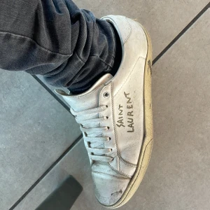 Vita Saint Laurent sneakers i skinn - Säljer ett par klassiska vita sneakers från Saint Laurent med handskriven logga på sidan. Skorna har rund tå, snörning och är tillverkade i skinn. Perfekta för dig som gillar stilrena och exklusiva sneakers med en urban vibe.